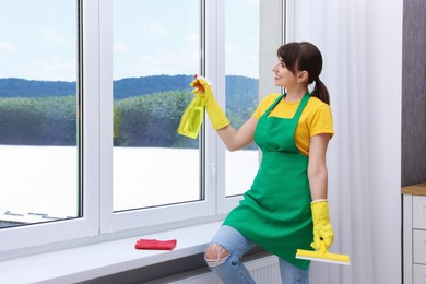 Photo of Housewife with squeegee and spray bottle of detergent cleaning window indoors