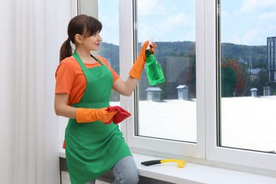 Housewife with spray bottle of detergent and napkin cleaning window indoors