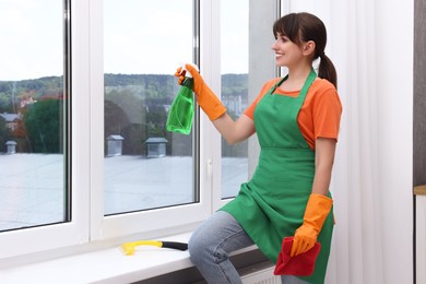 Housewife with spray bottle of detergent and napkin cleaning window indoors