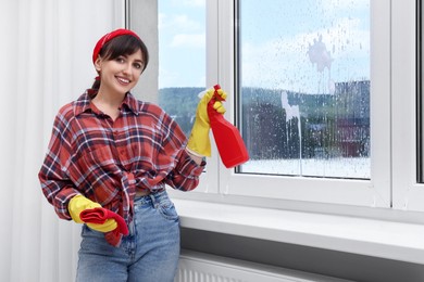 Beautiful young woman with spray bottle of detergent and napkin cleaning window indoors