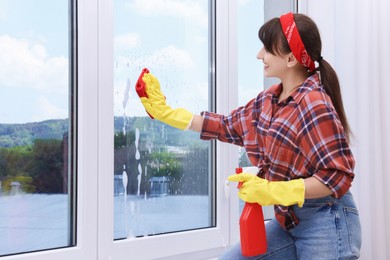 Photo of Beautiful young woman with spray bottle of detergent and napkin cleaning window indoors