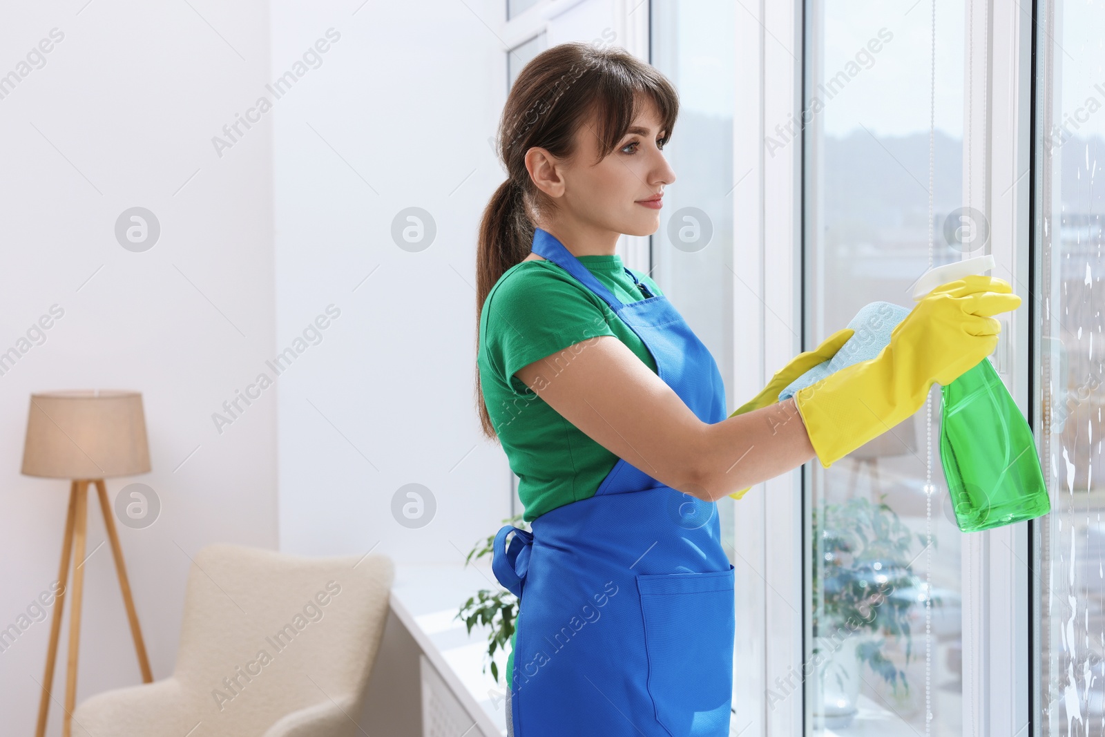 Photo of Beautiful young woman with spray bottle of detergent and napkin cleaning window indoors