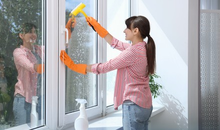 Beautiful young housewife with squeegee cleaning window indoors