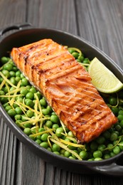 Photo of Delicious grilled salmon fillet with green peas and lime in baking dish on wooden table, closeup