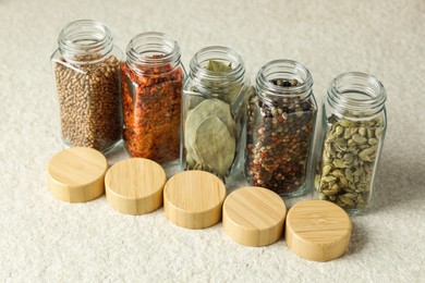 Photo of Different spices in glass jars and lids on light textured table, closeup