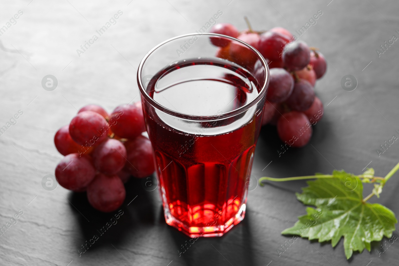 Photo of Tasty grape juice in glass, leaf and berries on dark textured table, closeup