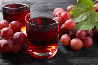 Tasty grape juice in glasses, leaf and berries on black wooden table, closeup