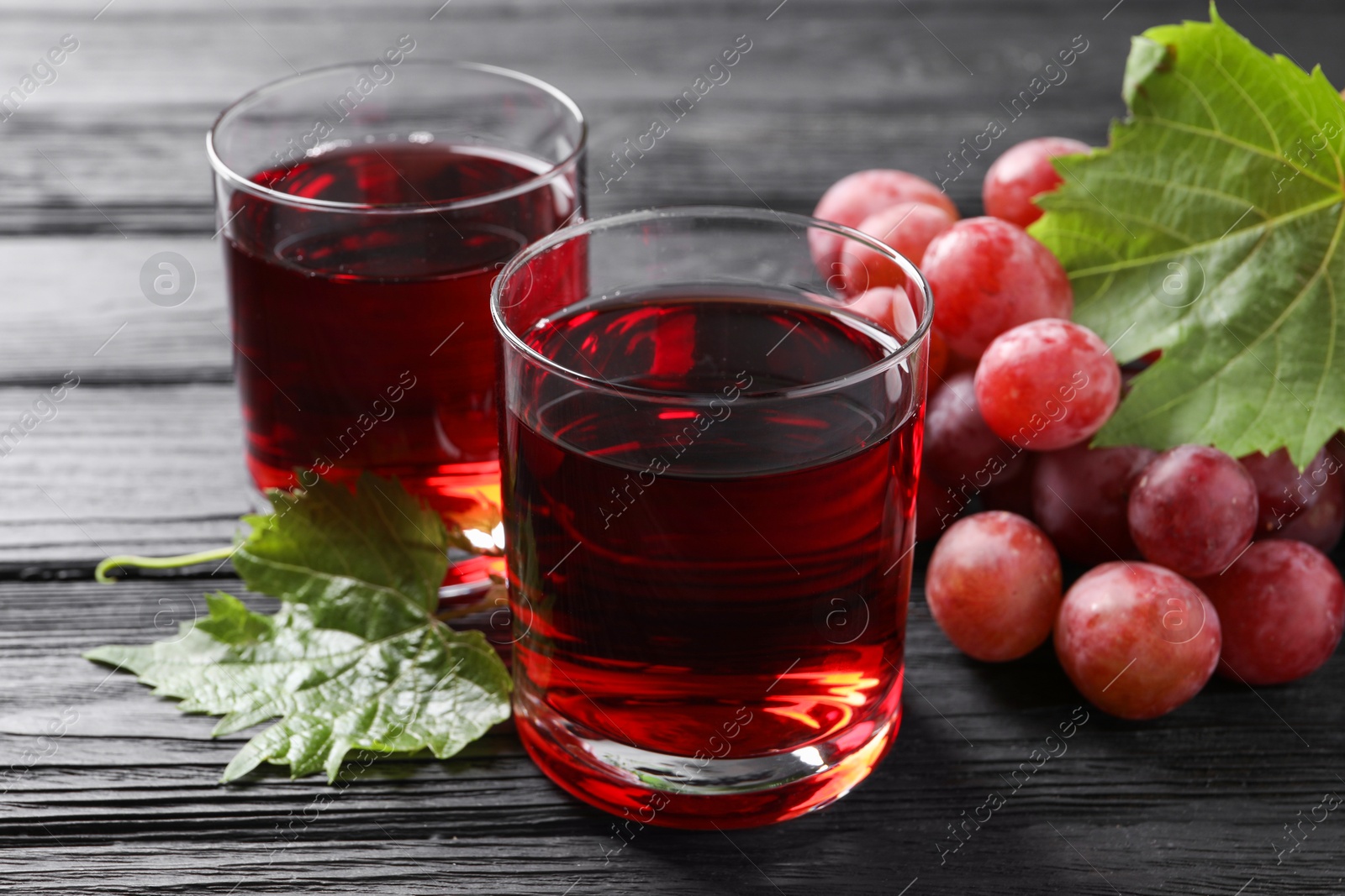 Photo of Tasty grape juice in glasses, leaves and berries on black wooden table, closeup