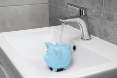 Photo of Water saving concept. Stream of water flowing from tap into piggy bank in bathroom