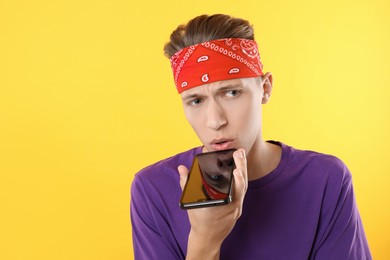 Young man recording voice message via smartphone on yellow background, space for text