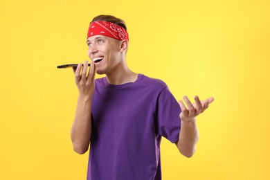 Young man recording voice message via smartphone on yellow background