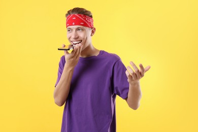 Young man recording voice message via smartphone on yellow background