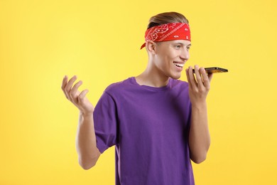 Young man recording voice message via smartphone on yellow background