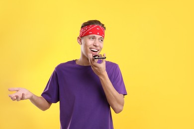 Young man recording voice message via smartphone on yellow background, space for text