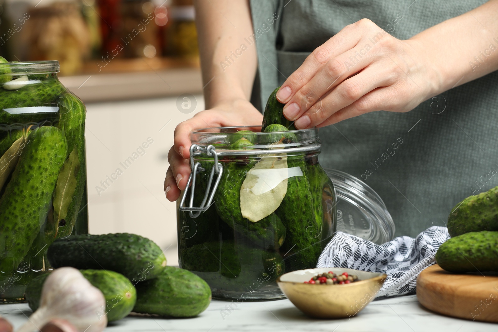 Photo of Making pickles. Woman putting cucumber into glass jar at white marble table, closeup