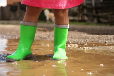 Photo of Little girl wearing green rubber boots standing in puddle outdoors, closeup