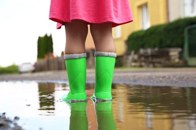 Photo of Little girl wearing green rubber boots standing in puddle outdoors, closeup