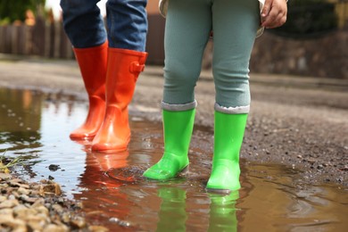 Photo of Mother and daughter wearing rubber boots standing in puddle outdoors, closeup