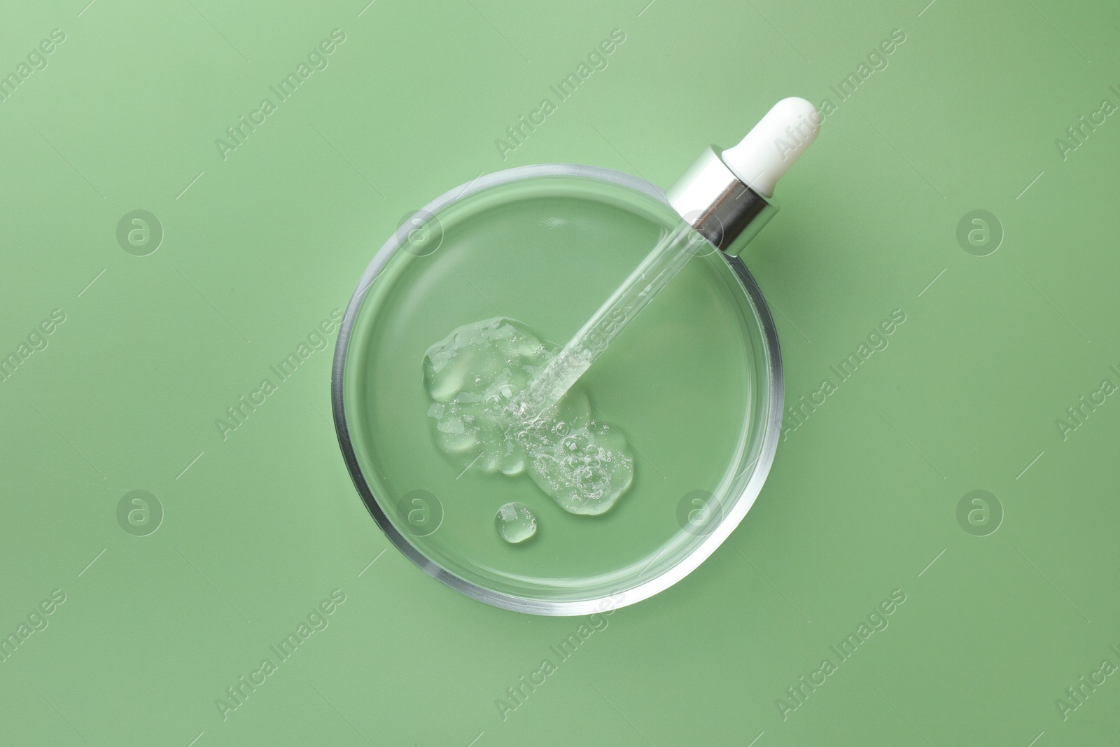 Photo of Petri dish with cosmetic product and pipette on green background, top view