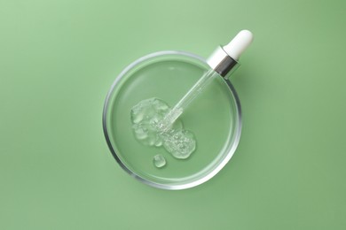 Photo of Petri dish with cosmetic product and pipette on green background, top view