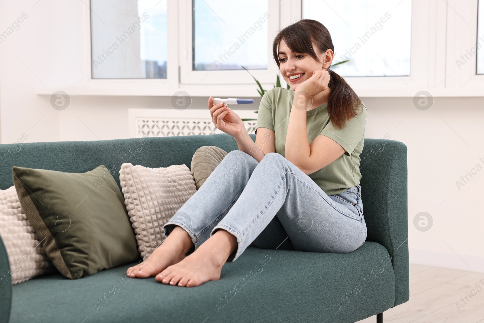 Photo of Happy young woman with pregnancy test on sofa at home