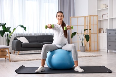 Beautiful pregnant woman with dumbbells doing exercises on fitball at home