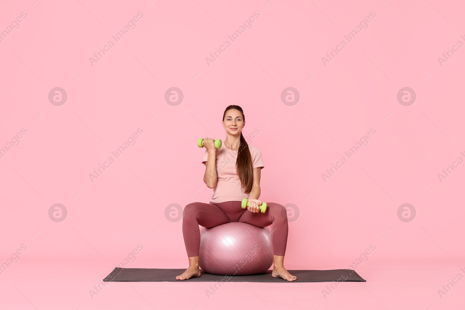 Photo of Beautiful pregnant woman with dumbbells doing exercises on fitball against pink background