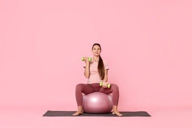 Photo of Beautiful pregnant woman with dumbbells doing exercises on fitball against pink background