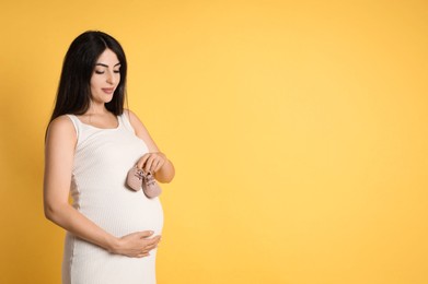 Photo of Portrait of beautiful pregnant woman with baby shoes on yellow background, space for text