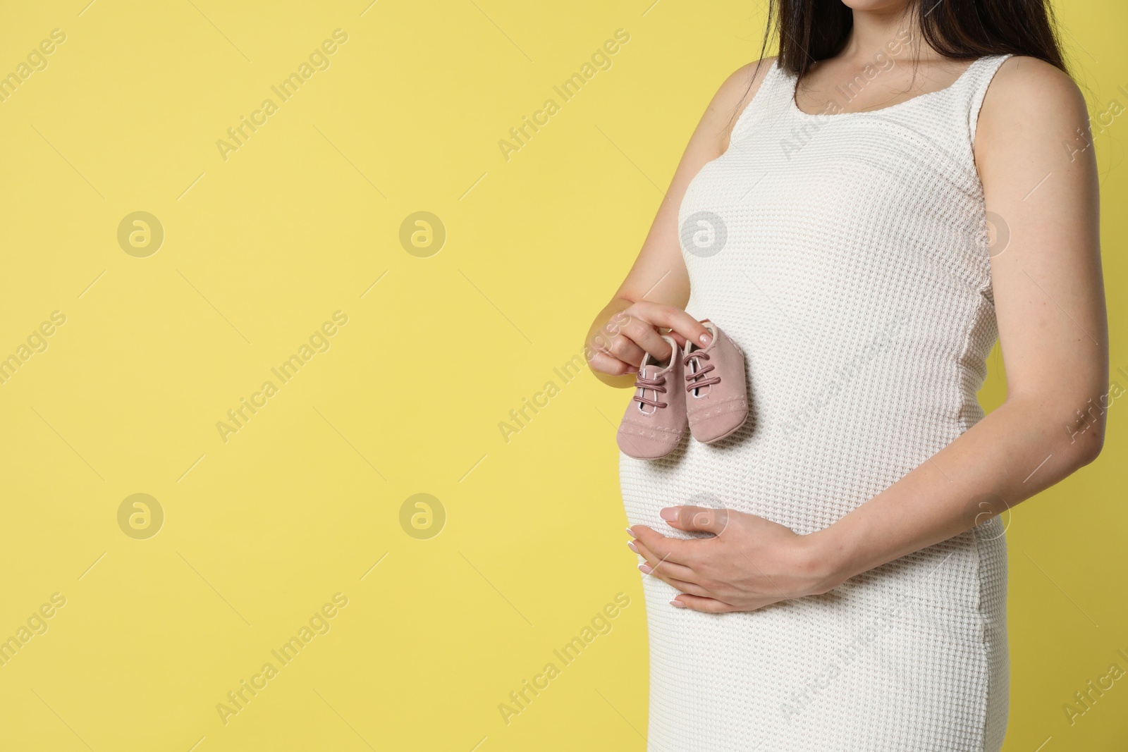 Photo of Young pregnant woman with pair of baby shoes on yellow background, closeup. Space for text