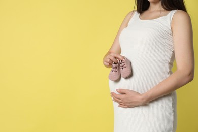 Photo of Young pregnant woman with pair of baby shoes on yellow background, closeup. Space for text