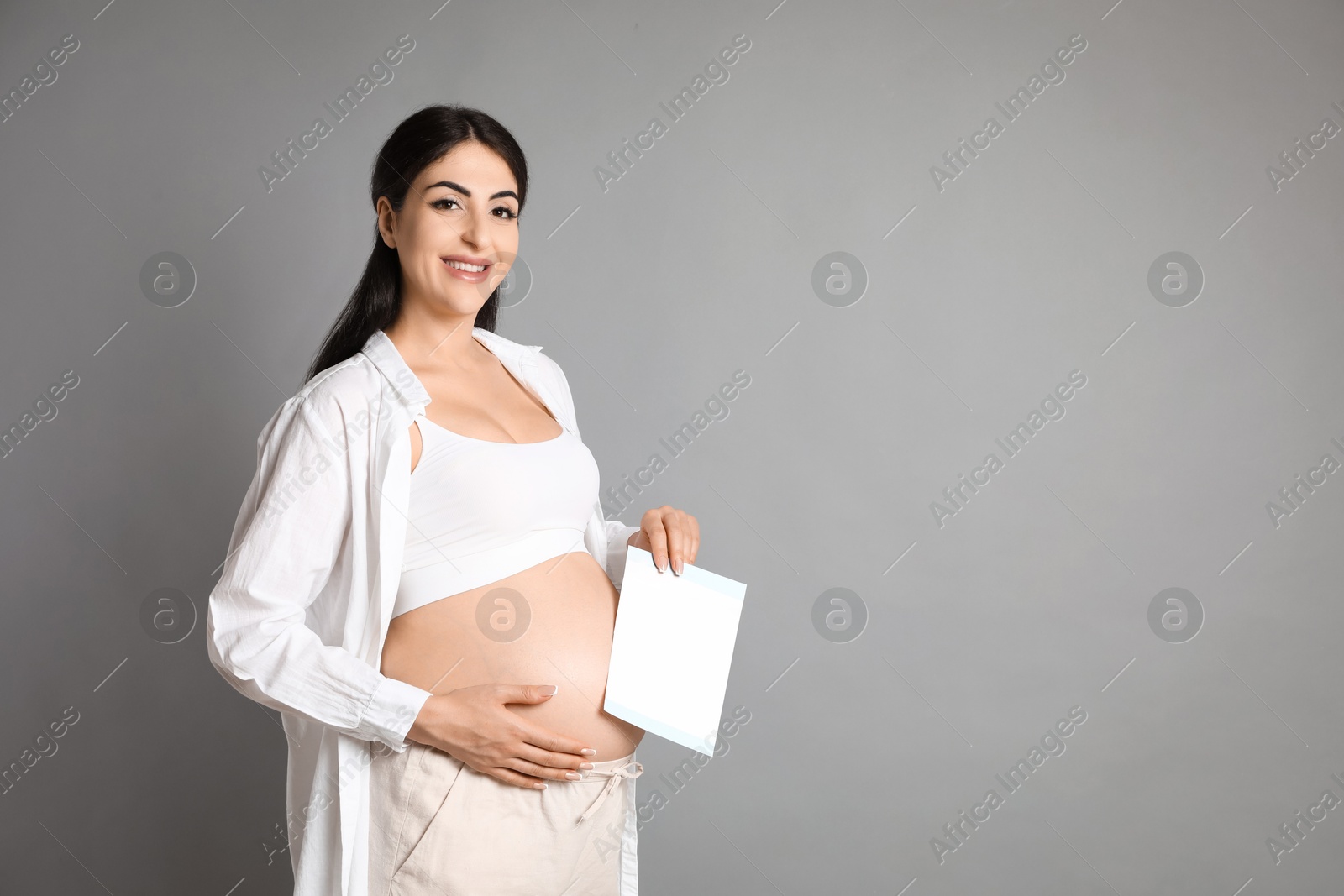 Photo of Portrait of beautiful pregnant woman with paper sheet on grey background, space for text