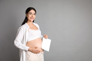 Portrait of beautiful pregnant woman with paper sheet on grey background, space for text