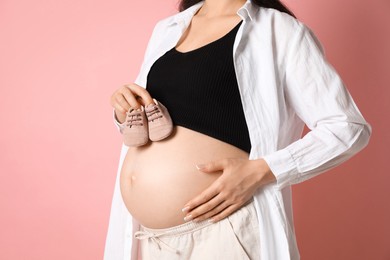 Photo of Young pregnant woman with pair of baby shoes on pink background, closeup