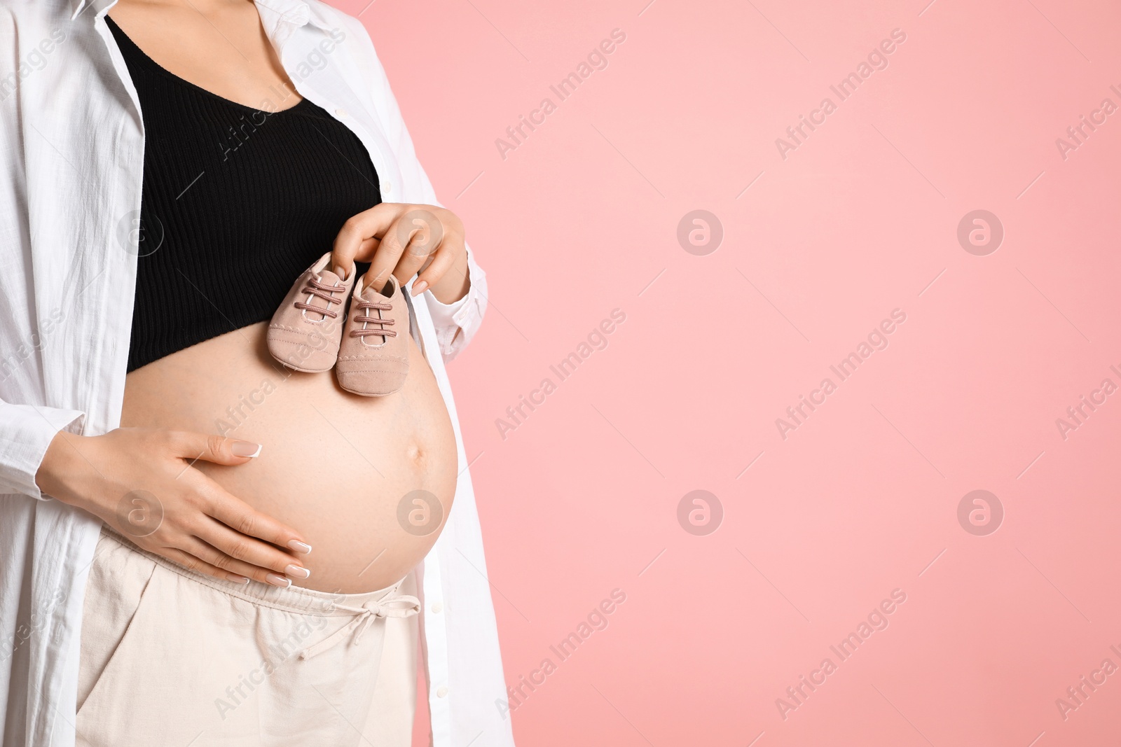 Photo of Young pregnant woman with pair of baby shoes on pink background, closeup. Space for text