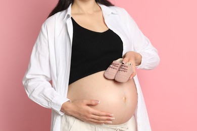 Young pregnant woman with pair of baby shoes on pink background, closeup