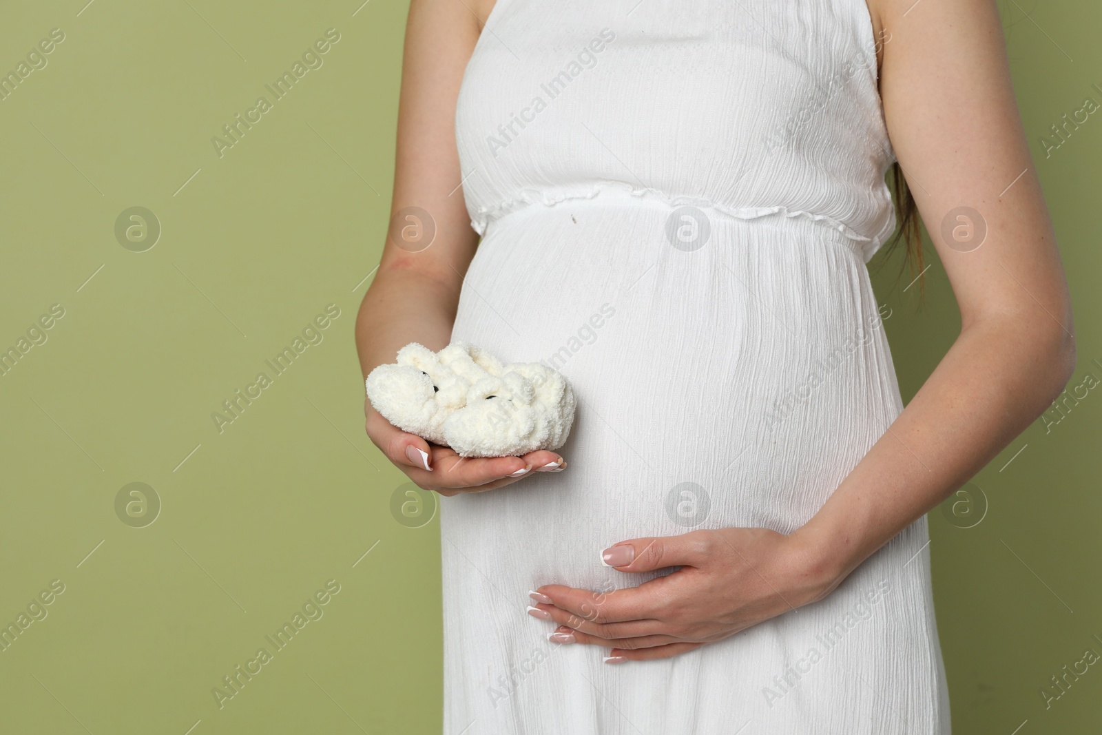 Photo of Young pregnant woman with pair of baby shoes on light olive background, closeup