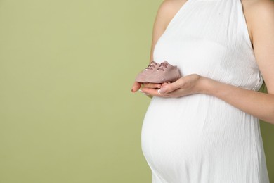 Photo of Young pregnant woman with pair of baby shoes on light olive background, closeup. Space for text