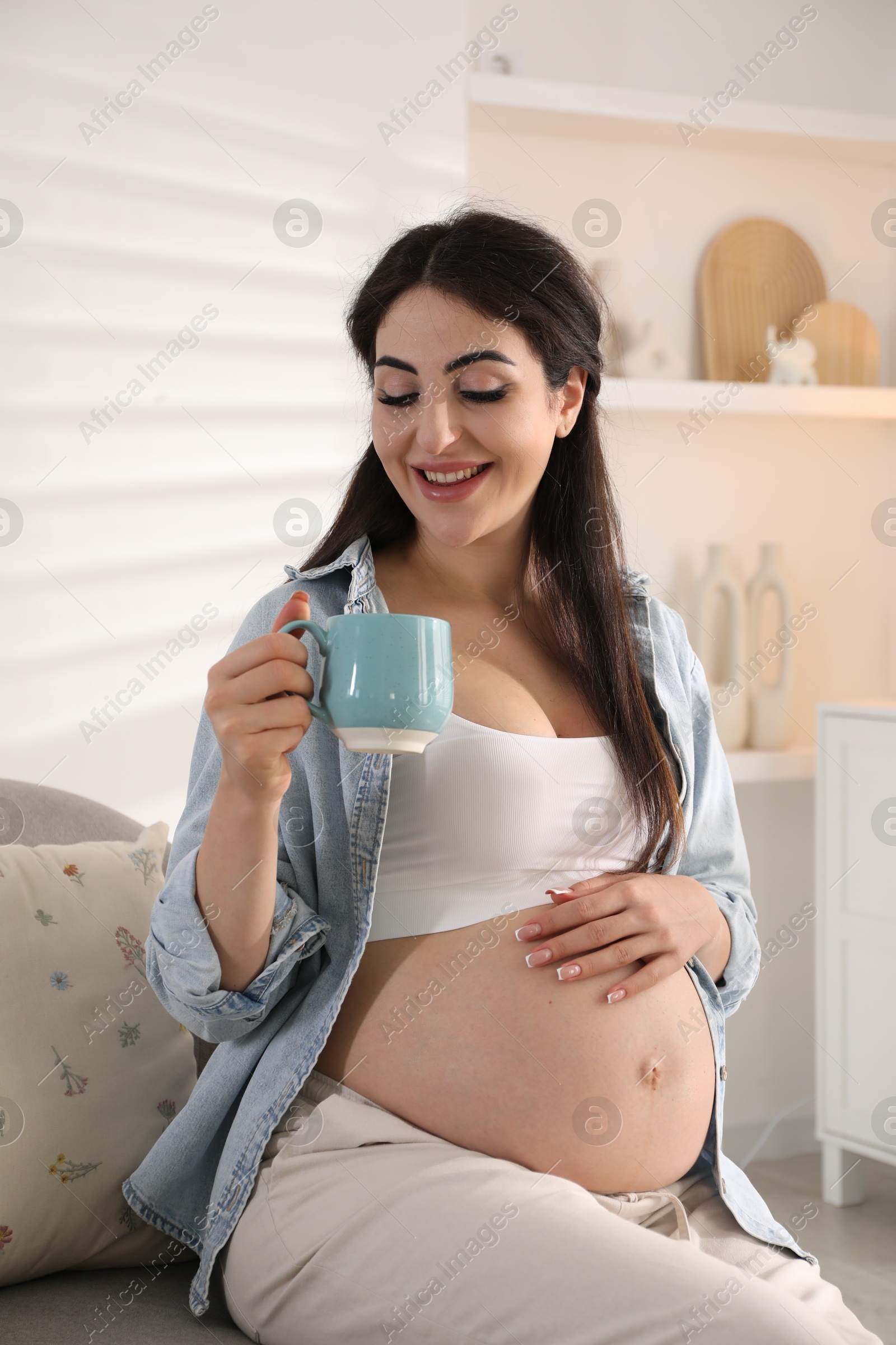 Photo of Young pregnant woman with cup of drink sitting on sofa at home