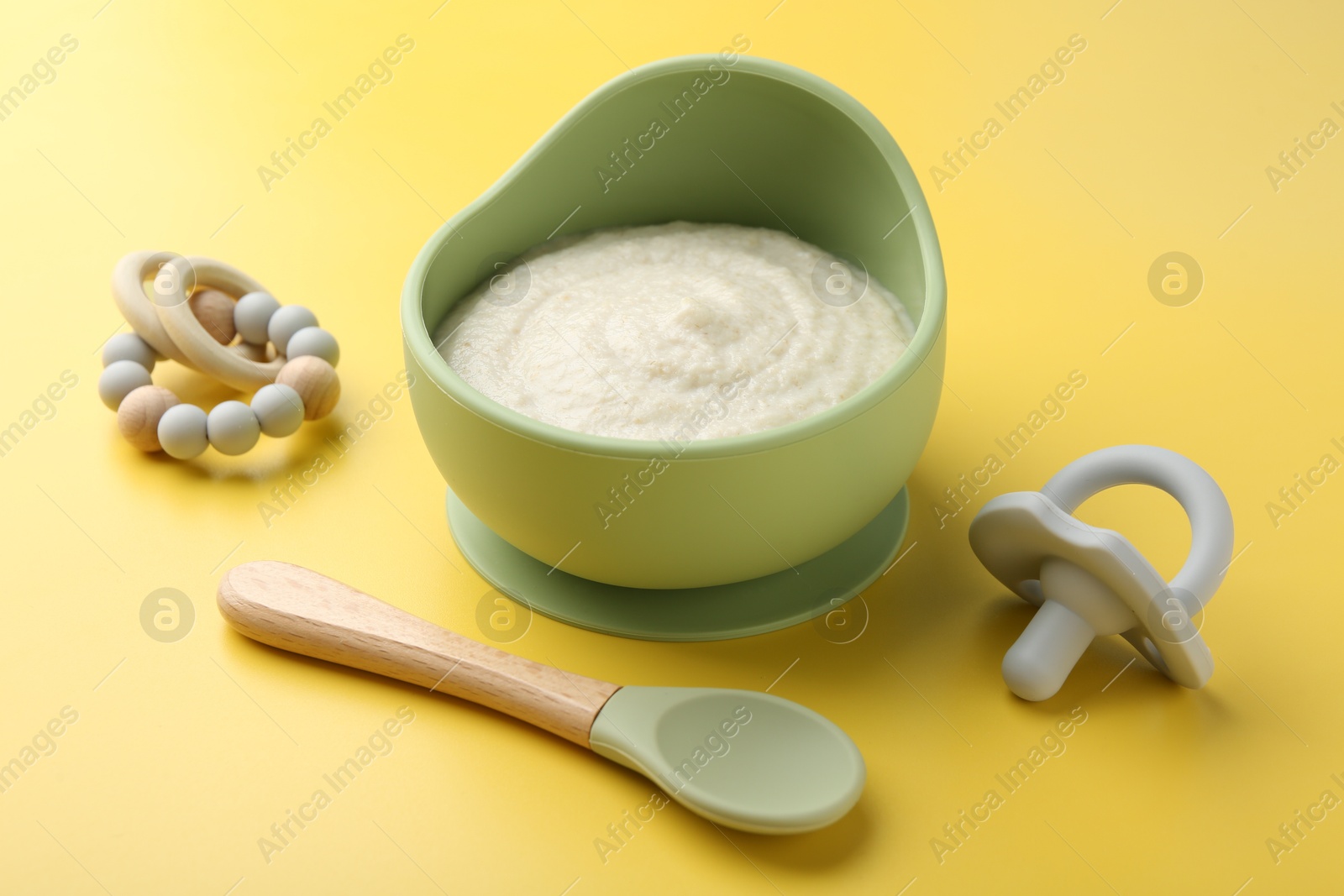 Photo of Delicious baby food in bowl with spoon, pacifier and teether on yellow background