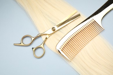 Blonde hair strand, comb and professional scissors on light grey background, top view