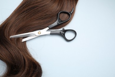 Photo of Brown hair strand and professional scissors on light grey background, top view. Space for text