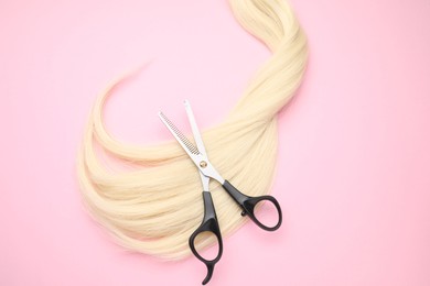 Photo of Blonde hair strand and professional scissors on pink background, top view