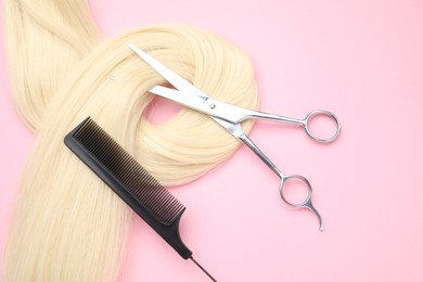 Blonde hair strand, comb and professional scissors on pink background, top view