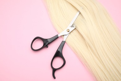 Blonde hair strand and professional scissors on pink background, top view