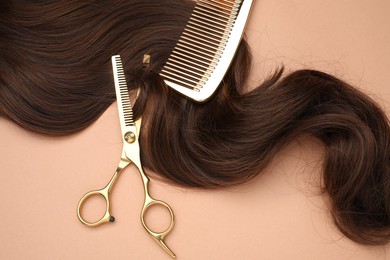 Brown hair strand, comb and professional scissors on pale coral background, top view