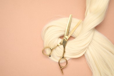 Photo of Blonde hair strand and professional scissors on pale coral background, top view. Space for text