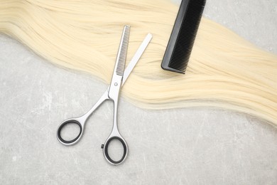 Photo of Blonde hair strand, comb and professional scissors on grey table, top view