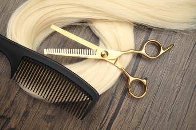 Photo of Blonde hair strand, comb and professional scissors on wooden surface, flat lay