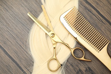 Blonde hair strand, comb and professional scissors on wooden surface, flat lay